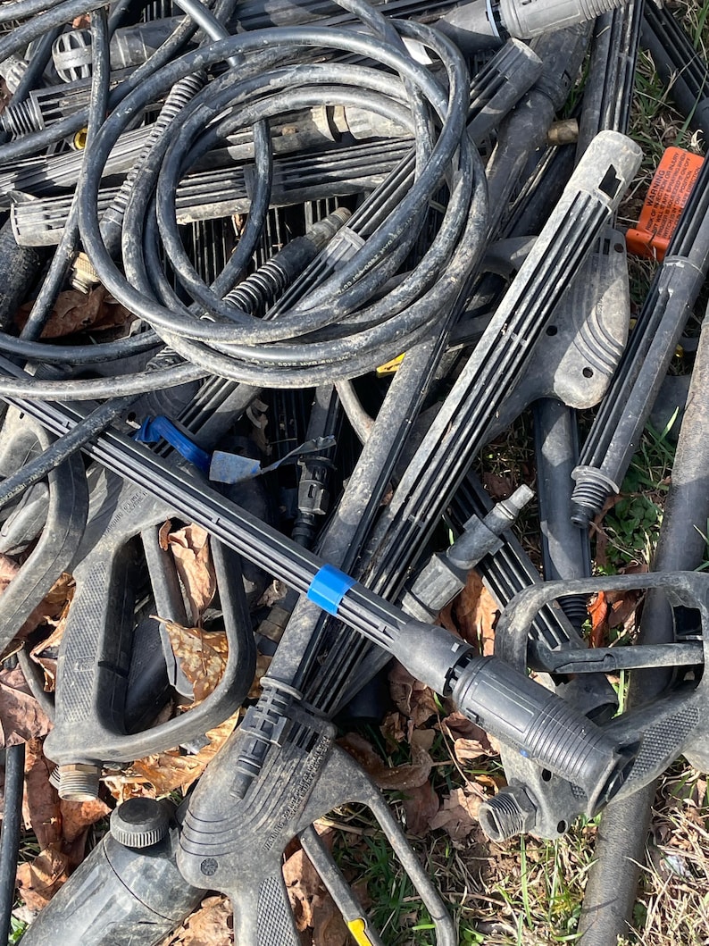 May include: A pile of black pressure washer parts, including hoses, spray wands, and handles. The components are scattered, with some showing signs of wear. A blue band is visible on one of the spray wands.
