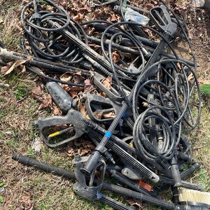 May include: A pile of black pressure washer parts, including hoses, spray wands, and handles, lies on the ground. The components are tangled together, with some showing signs of wear. A brush attachment is visible. The scene is set outdoors on grass and fallen leaves.