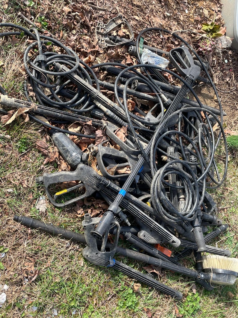 May include: A pile of black pressure washer parts, including hoses, spray wands, and handles, lies on the ground. The components are tangled together, with some showing signs of wear. A brush attachment is visible. The scene is set outdoors on grass and fallen leaves.