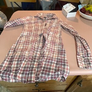 Op de afbeelding: Een geruit shirt met lange mouwen in rood-, wit- en bruintinten, met knopen aan de voorkant. Het shirt ligt plat op een roze ondergrond, met de mouwen uitgestrekt. Het shirt heeft een klassieke kraag en manchetten met knopen.