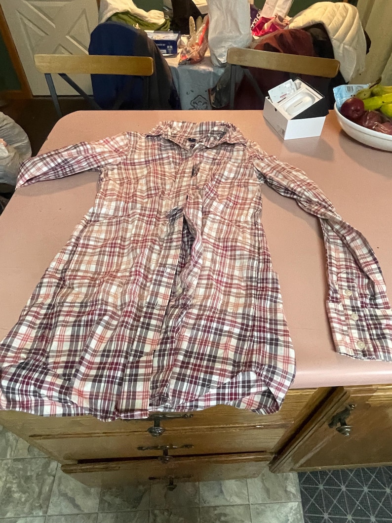 Op de afbeelding: Een geruit shirt met lange mouwen in rood-, wit- en bruintinten, met knopen aan de voorkant. Het shirt ligt plat op een roze ondergrond, met de mouwen uitgestrekt. Het shirt heeft een klassieke kraag en manchetten met knopen.