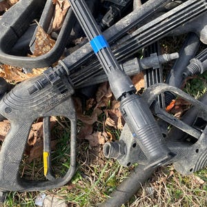 May include: A pile of black pressure washer parts, including handles, wands, and nozzles. Some parts have yellow and blue accents. The items are lying on grass and surrounded by fallen leaves.