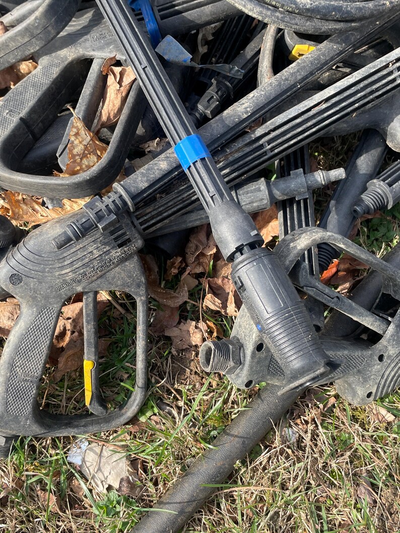 May include: A pile of black pressure washer parts, including handles, wands, and nozzles. Some parts have yellow and blue accents. The items are lying on grass and surrounded by fallen leaves.