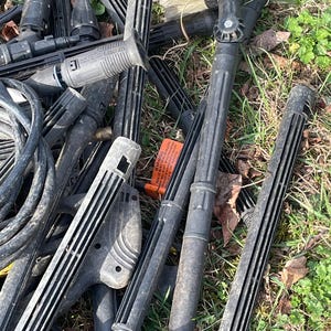 May include: A collection of black pressure washer parts, including wands, nozzles, and brushes, scattered on a bed of dry leaves and grass. The parts vary in size and shape, with some featuring ribbed designs and others with brush heads.