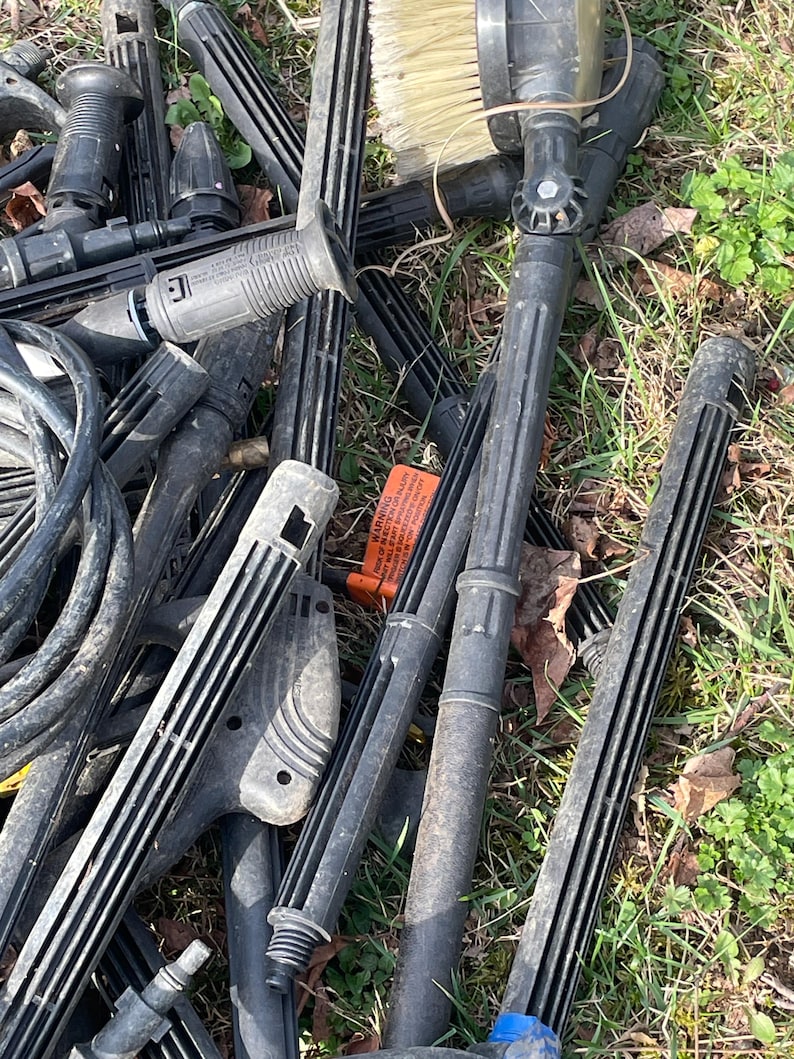 May include: A collection of black pressure washer parts, including wands, nozzles, and brushes, scattered on a bed of dry leaves and grass. The parts vary in size and shape, with some featuring ribbed designs and others with brush heads.