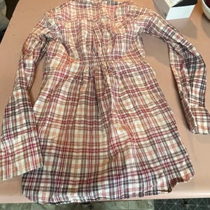 Op de afbeelding: Een lange mouwen plaid jurk in tinten rood, bruin en cr&egrave;me. De jurk heeft een getailleerde taille en een uitlopende rok. De stof heeft een klassiek ruitpatroon en de mouwen hebben manchetten met knopen. De jurk ligt op een lichtroze ondergrond.