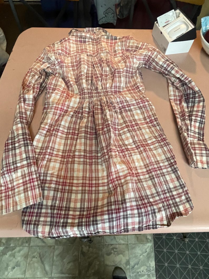 Op de afbeelding: Een lange mouwen plaid jurk in tinten rood, bruin en cr&egrave;me. De jurk heeft een getailleerde taille en een uitlopende rok. De stof heeft een klassiek ruitpatroon en de mouwen hebben manchetten met knopen. De jurk ligt op een lichtroze ondergrond.