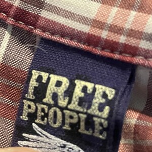 Op de afbeelding: Detail van een geruit overhemd met een label met de tekst "FREE PEOPLE" in een verweerde, gebroken witte letter. De stof van het overhemd heeft een patroon van rode, witte en roze vierkanten.