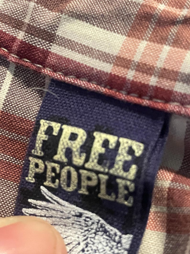 Op de afbeelding: Detail van een geruit overhemd met een label met de tekst "FREE PEOPLE" in een verweerde, gebroken witte letter. De stof van het overhemd heeft een patroon van rode, witte en roze vierkanten.