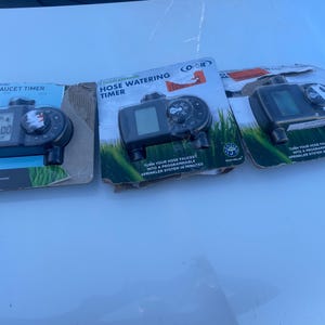 May include: Three black hose faucet timers in their original packaging. Each timer has a digital display and a dial for setting the watering schedule. The packaging features text and graphics, including the words "Hose Faucet Timer" and "Hose Watering Timer."