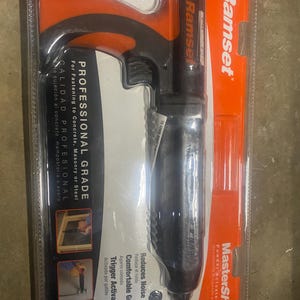 May include: A Ramset MasterShot powder-actuated tool, designed for professional use. The tool is black and orange, with the Ramset logo visible. Features include a comfortable grip and trigger activation. Text on the packaging indicates it's for fastening to concrete, masonry, or steel.