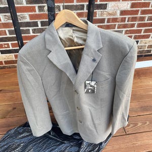 May include: A light gray blazer with a two-button closure, hanging on a wooden hanger. The blazer has a notched lapel, a small pin, and a tag attached to the pocket. The background features a brick wall and a black surface.