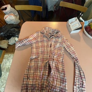 Op de afbeelding: Een overhemd met lange mouwen en knopen in een ruitpatroon van rood, wit en beige. Het overhemd ligt plat op een roze ondergrond, met de mouwen uitgestrekt. De kraag is zichtbaar en het overhemd lijkt gemaakt van een lichte stof.