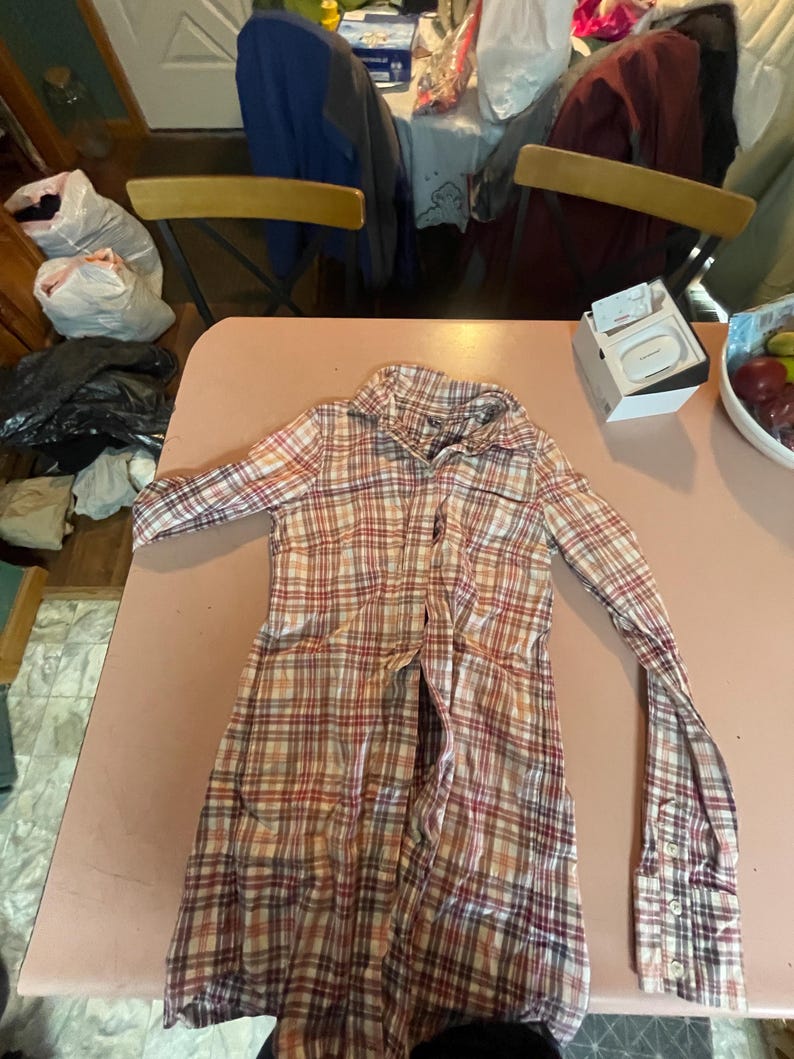 Op de afbeelding: Een overhemd met lange mouwen en knopen in een ruitpatroon van rood, wit en beige. Het overhemd ligt plat op een roze ondergrond, met de mouwen uitgestrekt. De kraag is zichtbaar en het overhemd lijkt gemaakt van een lichte stof.