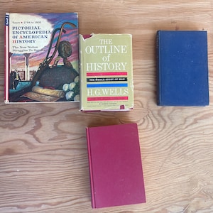 May include: Four vintage books are arranged on a wooden surface. The books include a pictorial encyclopedia of American history, a book titled "The Outline of History" by H.G. Wells, and two other books with solid-colored covers in blue and red.