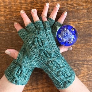 May include: Teal fingerless gloves with a cable knit design. A blue glass sphere with embedded bubbles rests nearby. The gloves are made of a soft yarn and feature a textured pattern.