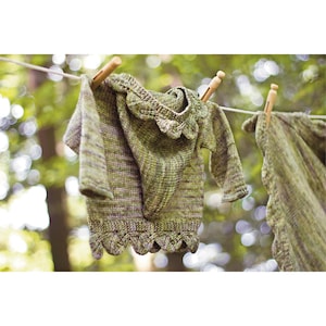 May include: A green knitted sweater and shawl are hanging on a clothesline with wooden clothespins. The sweater has a scalloped edge and long sleeves. The shawl also has a scalloped edge. The background is a blurred view of green foliage.