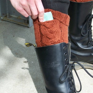 May include: Rust-colored cable knit leg warmers with a pocket, worn over black pants and black combat boots. The boots have black laces and a yellow tag. A hand is placing a card into the pocket.