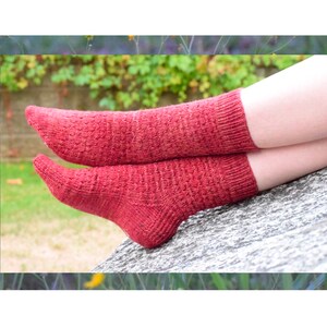 May include: A pair of red knitted socks with a textured pattern. The socks are worn on a person's feet and are resting on a stone surface.