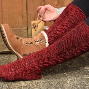 May include: A pair of red knitted leg warmers with a cable knit pattern. The leg warmers are worn over brown boots.