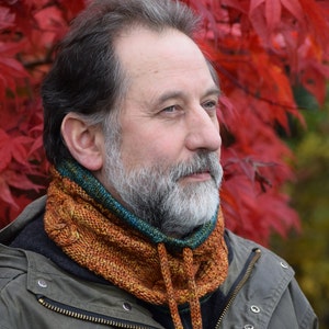 Puede incluir: Un hombre con barba gris que lleva una bufanda de punto marrón, verde y naranja. La bufanda tiene un cordón largo que se puede atar. El hombre lleva una chaqueta verde oscura.