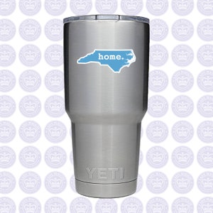 May include: A stainless steel Yeti tumbler with a light blue sticker of the North Carolina state outline and the word "home." The tumbler is cylindrical and has a silver lid. The Yeti logo is at the bottom.