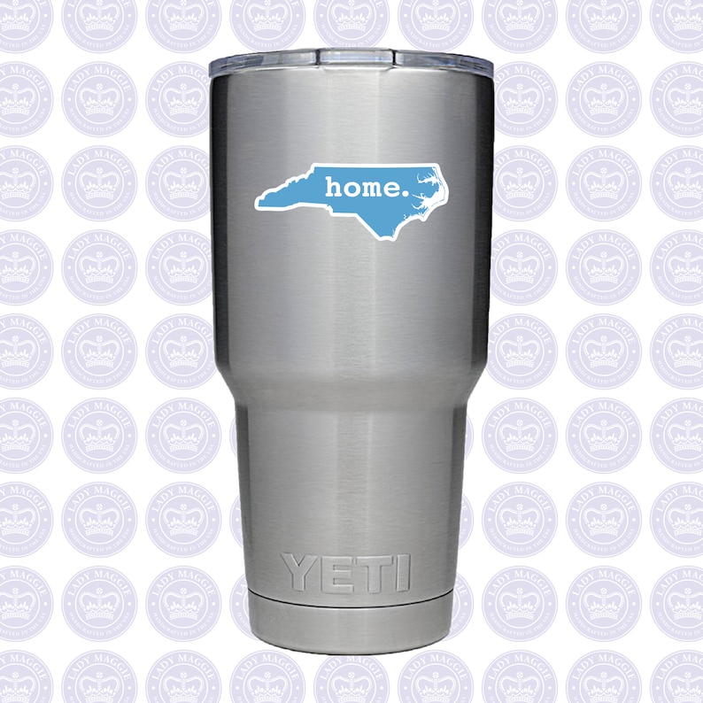May include: A stainless steel Yeti tumbler with a light blue sticker of the North Carolina state outline and the word "home." The tumbler is cylindrical and has a silver lid. The Yeti logo is at the bottom.