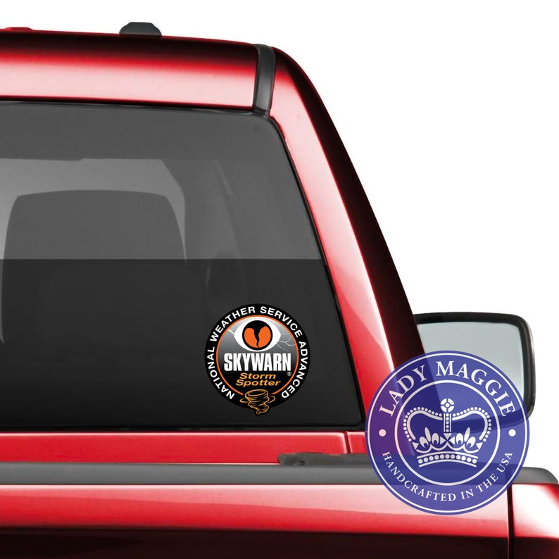 May include: A red pickup truck with a black window sticker that says "National Weather Service Advanced SKYWARN Storm Spotter". The sticker has a black and orange design with a stylized eye and lightning bolt. A blue and white sticker with a crown and the text "Lady Maggie Handcrafted in the USA" is on the truck's rear passenger side window.