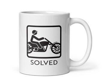 May include: White ceramic mug with a black silhouette of a person riding a motorcycle inside a square. The text "SOLVED" is printed below the image.