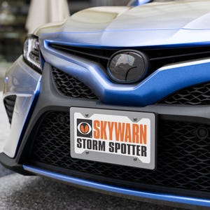 May include: A license plate with the text "SKYWARN STORM SPOTTER" in orange and black lettering. The plate is mounted on the front of a blue car.