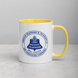 May include: White ceramic mug with a yellow rim and handle. The mug features a blue and white logo of the American Telephone & Telegraph Co. with the text "Local and Long Distance Telephone Bell System and Associated Companies".