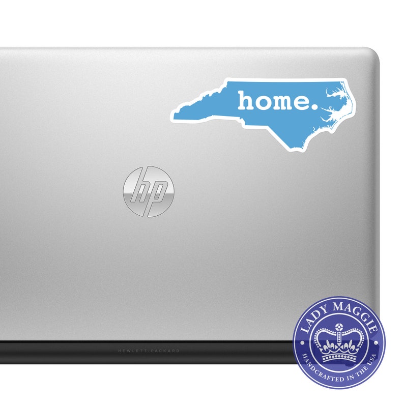 May include: A light blue sticker in the shape of North Carolina with the word "home." in white text, affixed to a silver laptop. A circular sticker with a crown and the text "Lady Maggie Handcrafted in the USA" is in the lower right corner.
