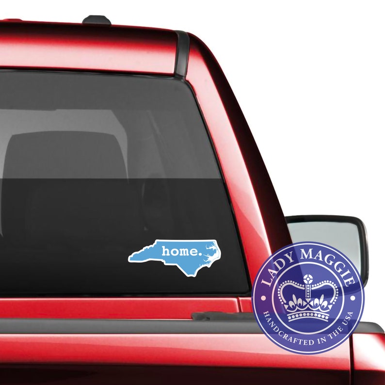May include: A light blue sticker in the shape of North Carolina with the word "home" in a white font. The sticker is on the rear window of a red vehicle. A circular logo with a crown and the words "Lady Maggie" is also visible.