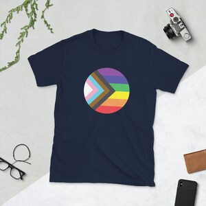 Puede incluir: Camiseta azul marino con un diseño circular que presenta una bandera arcoíris con un chevron negro y marrón en su interior. El chevron está coronado por un triángulo blanco y azul claro.