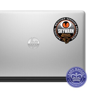 May include: A silver laptop computer with a black HP logo and two stickers. The first sticker is a black and orange circle with the text "National Weather Service Advanced SKYWARN Storm Spotter" and a stylized image of a tornado. The second sticker is a blue circle with the text "Lady Maggie Handcrafted in the USA" and a crown.