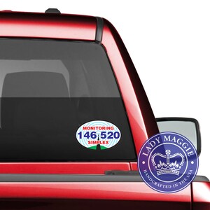 May include: A red pickup truck with two decals on the back window. The left decal is a blue oval with white text that reads "Monitoring 146.520 Simplex". The right decal is a blue circle with a crown and the text "Lady Maggie Handcrafted in the USA".