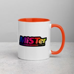 May include: White ceramic mug with an orange rim and handle. The mug features a colorful logo that says "MiSTer FPGA".