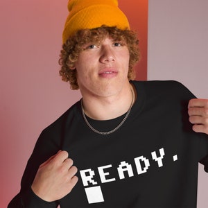 Könnte beinhalten: Ein schwarzes Sweatshirt mit einer weißen pixelartigen Grafik, auf der "READY." steht. Die Grafik enthält auch ein weißes Quadrat. Die Person, die das Sweatshirt trägt, hat lockiges rotes Haar und trägt eine gelbe Mütze.