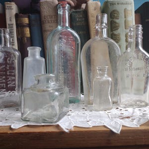 May include: A collection of antique glass bottles of various shapes and sizes, some with embossed text. The bottles are clear and arranged on a wooden surface in front of a backdrop of old books. The bottles vary in height.