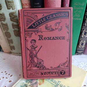 Könnte beinhalten: Ein rotes Vintage-Buch "Little Classics Romance", Band 7, mit schwarzen dekorativen Elementen und Text. Weitere antike Bücher sind sichtbar, die ein klassisches literarisches Thema schaffen. Das Buch ist von anderen antiken Büchern umgeben.