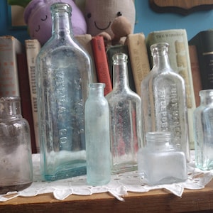 May include: Collection of antique glass bottles of various shapes and sizes, including a large aqua-colored bottle with text. The bottles are displayed on a wooden surface with a lace doily, against a backdrop of old books.