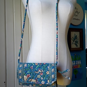 May include: A blue crossbody bag with a floral and animal print, including elephants and rabbits. The bag has a long, patterned strap and a flap closure. The background is a white mannequin and a blue wall.