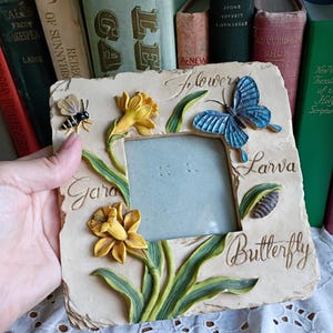 May include: A square beige picture frame with a textured stone-like finish. The frame features raised, painted decorations including yellow daffodils, green leaves, a small bee, a blue butterfly, and a chrysalis. The words Flowers, Larva, and Butterfly are embossed on the surface.