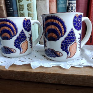 Puede incluir: Dos tazas de cerámica blanca con diseños florales azules y naranjas. Las tazas tienen un asa en el lado derecho.