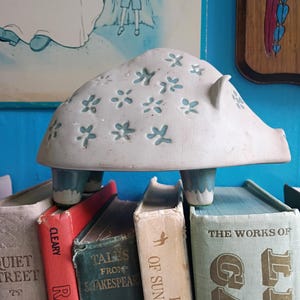 May include: A ceramic pig-shaped figurine with a matte off-white finish and a pattern of small, light blue, flower-like cutouts across its back and sides. The figurine stands on four short, sturdy legs that are glazed in a solid light blue color.