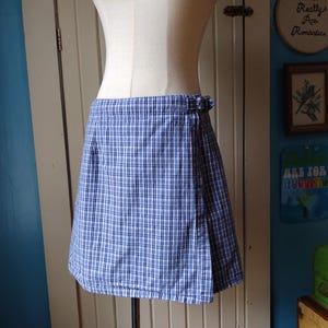 May include: A blue and white plaid wrap skirt displayed on a mannequin. The skirt is knee-length and features a classic plaid pattern. The fabric appears to be cotton or a similar material. The skirt is secured with a buckle.