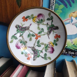 May include: A small, white porcelain dish with a gold rim. The dish features a floral and butterfly design in purple, green, yellow, and orange. The dish is placed on a stack of books.