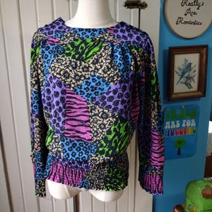 May include: A long-sleeved blouse with a patchwork design featuring animal prints in vibrant colours. The blouse has a round neckline, elasticated waist, and cuffs. Colours include purple, blue, green, black, and pink.