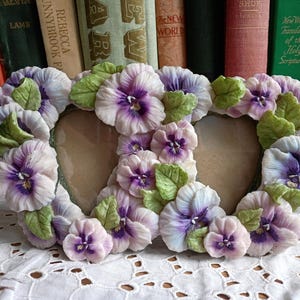 May include: A double heart-shaped picture frame decorated with sculpted pansy flowers in shades of light purple, white, and deep violet, accented with textured green leaves. The frame features two heart-shaped openings for photographs.