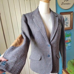 Puede incluir: Una blazer de tweed gris con coderas marrones y botones azules. La blazer tiene un diseño clásico con solapa de muesca y dos bolsillos delanteros. La chaqueta se muestra en un maniquí.